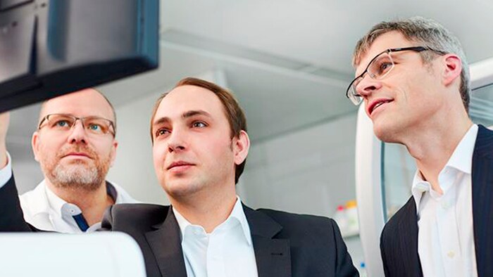 Three men — including two co-founders of Noscendo, Dr.  Philip Stevens and Dr. Peter Haug — look at a computer screen together. 